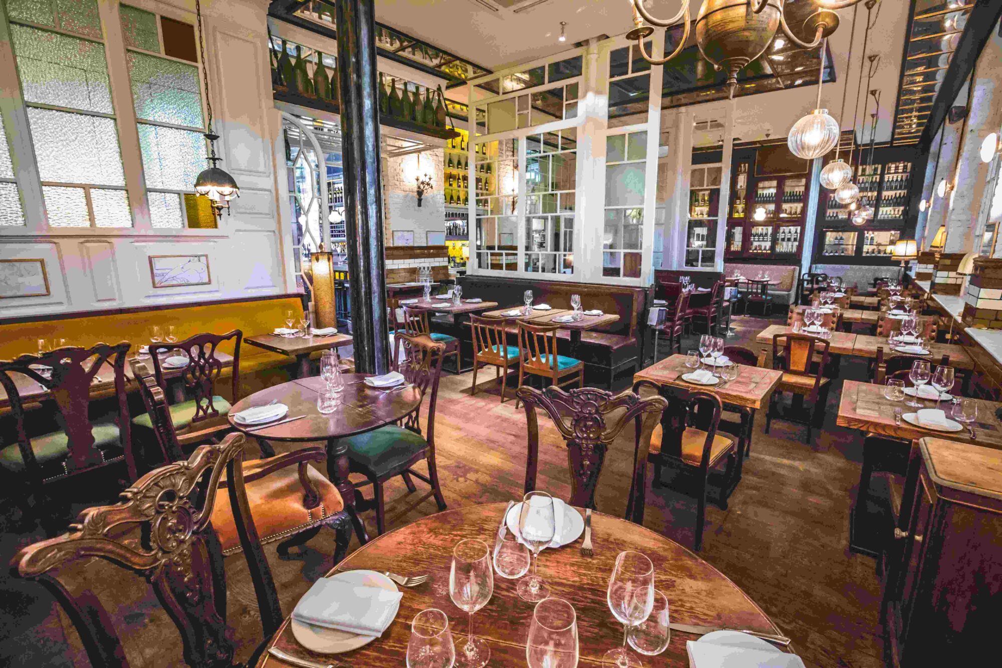 A cozy, elegant restaurant interior featuring wooden tables set with plates and glasses. Vintage chairs surround the tables, and large windows add light to the space. Decorative lighting and wine shelves enhance the warm atmosphere.