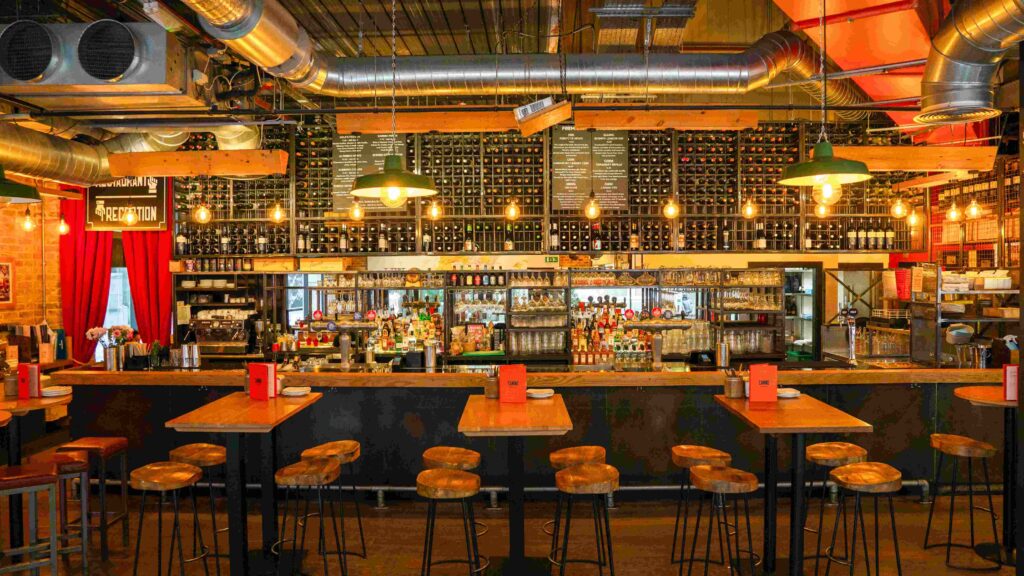 A cozy bar interior with wooden high tables and stools in the foreground. The back wall features shelves filled with wine bottles. Warm pendant lights hang from the ceiling, and a well-stocked bar area is in the center.