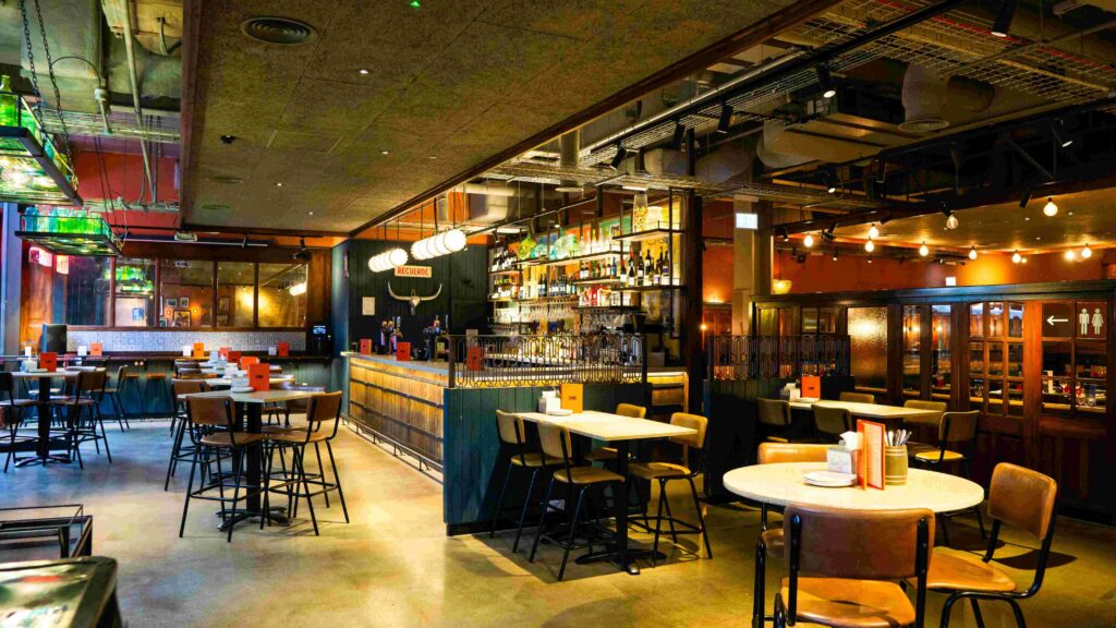 A spacious, stylish bar and restaurant interior with a mix of high tables, bar stools, and round tables with chairs. The bar is stocked with various bottles, and warm lighting creates a cozy atmosphere. Industrial-style ceilings are visible.