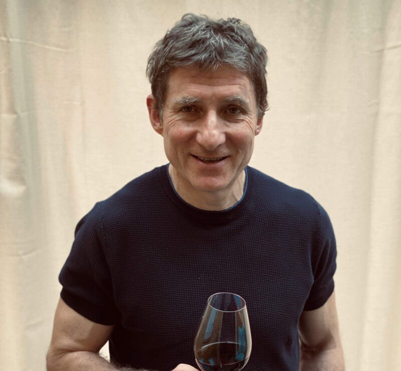 A person with short hair, wearing a dark shirt, is holding a glass of red wine and smiling. The background is a light-colored curtain.