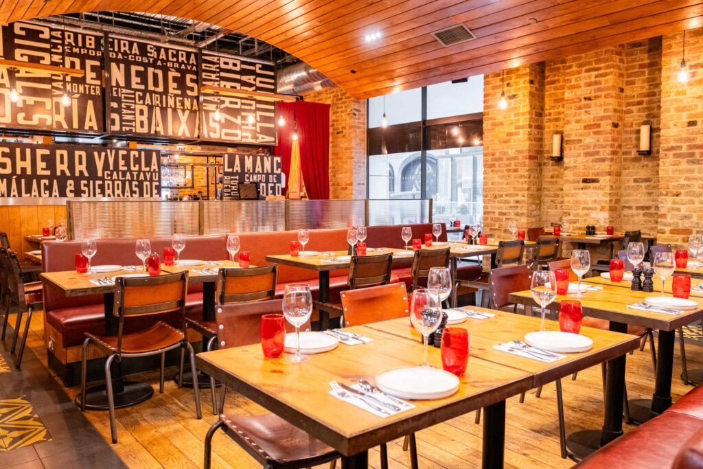 A cozy restaurant interior with wooden tables set for dining, featuring red glasses and brick walls. The decor includes large, bold text with various Spanish wine regions. Warm lighting and red curtains add to the inviting atmosphere.