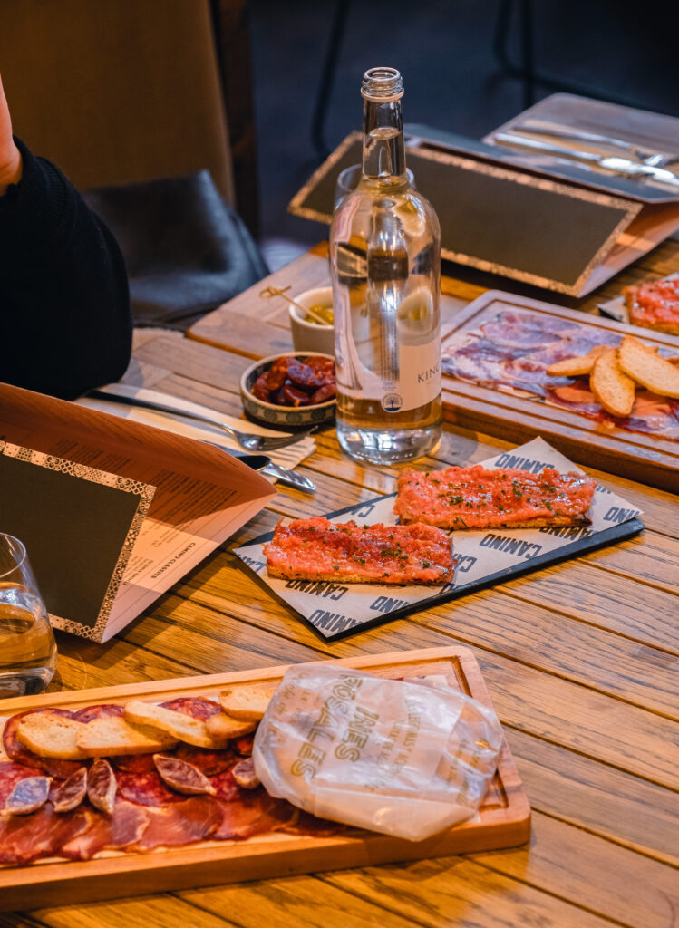 A wooden table with plates of sliced cured meats and bread, a bottle of water, and menus. There are small bowls of condiments, and a person partially visible holding a menu. Napkins and utensils are set beside the plates.