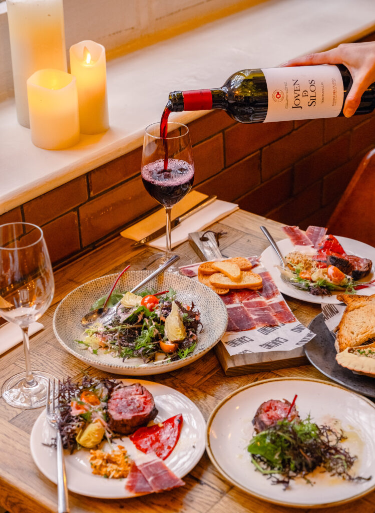 A cozy table setting with plates of gourmet appetizers, including salad and cured meats. A hand pours red wine into a glass. Lit candles create a warm ambiance. Napkins and cutlery are neatly arranged beside the dishes.