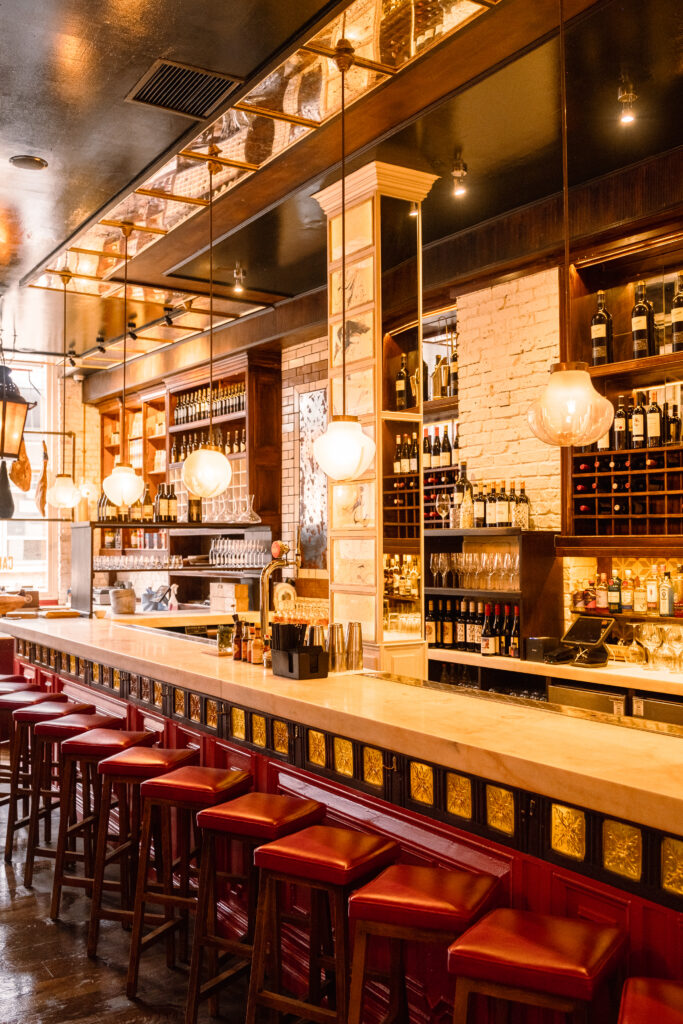 Cozy bar interior with a long marble counter and red cushioned barstools. Warm lighting from hanging globes illuminates shelves stocked with wine bottles and glasses. Decorative tiles adorn the bar, creating a welcoming atmosphere.