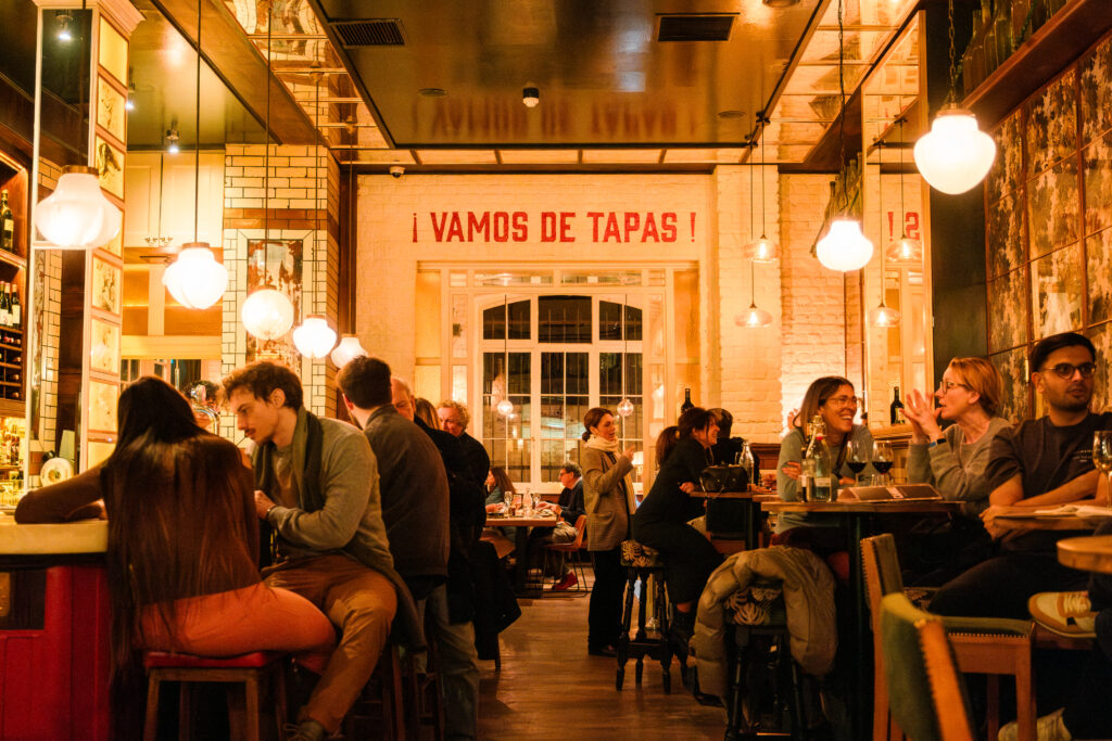 A lively tapas bar filled with people sitting and socializing at tables. Warm lighting from round hanging lamps creates a cozy atmosphere. A sign reads 