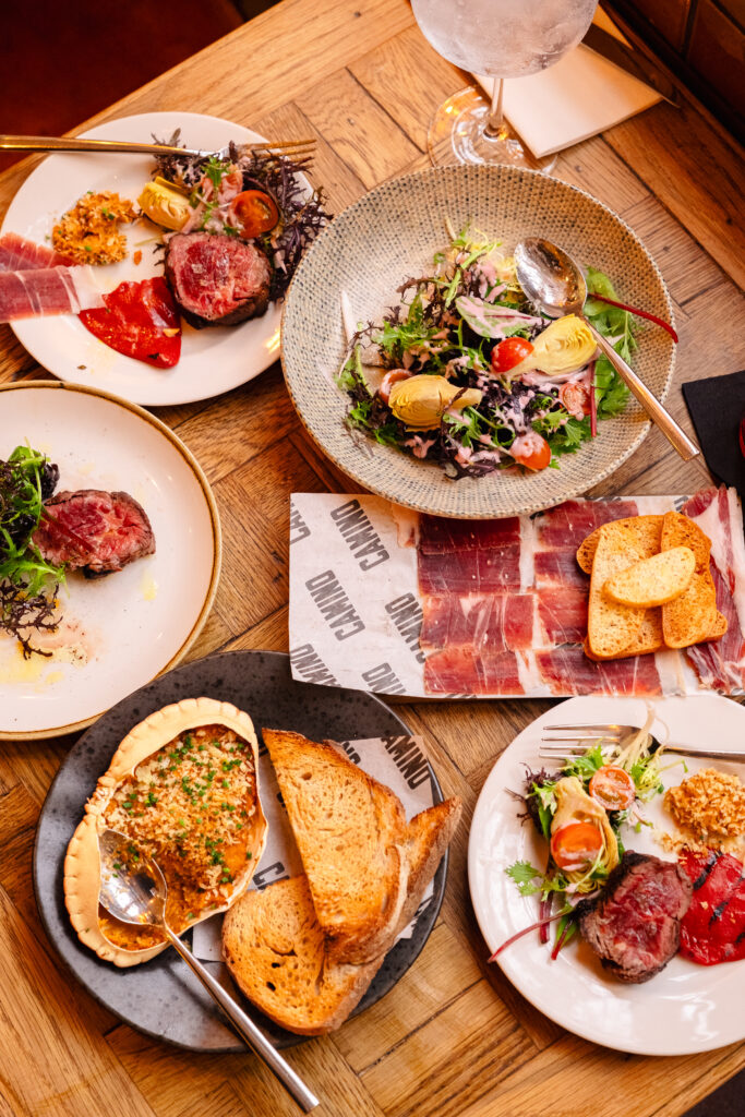 A wooden table is set with various plates of gourmet dishes, including salads, meat, and toasted bread. There's a plate of sliced prosciutto, a dish with stuffed vegetables, and a drink in a glass nearby. Plates are artfully arranged.