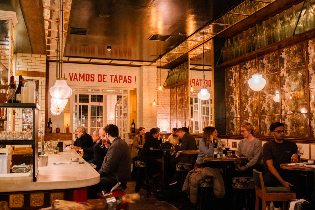 People dining in a cozy tapas bar with a warm atmosphere. The interior features wooden tables, hanging globe lights, and mirrored walls. A sign reading 