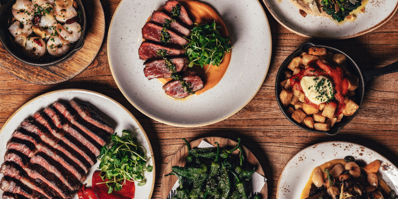 An overhead view of a wooden table with various dishes, including sliced steaks, roasted potatoes, prawns, blistered peppers, and a dish with beans. Each plate is artfully arranged with garnishes.