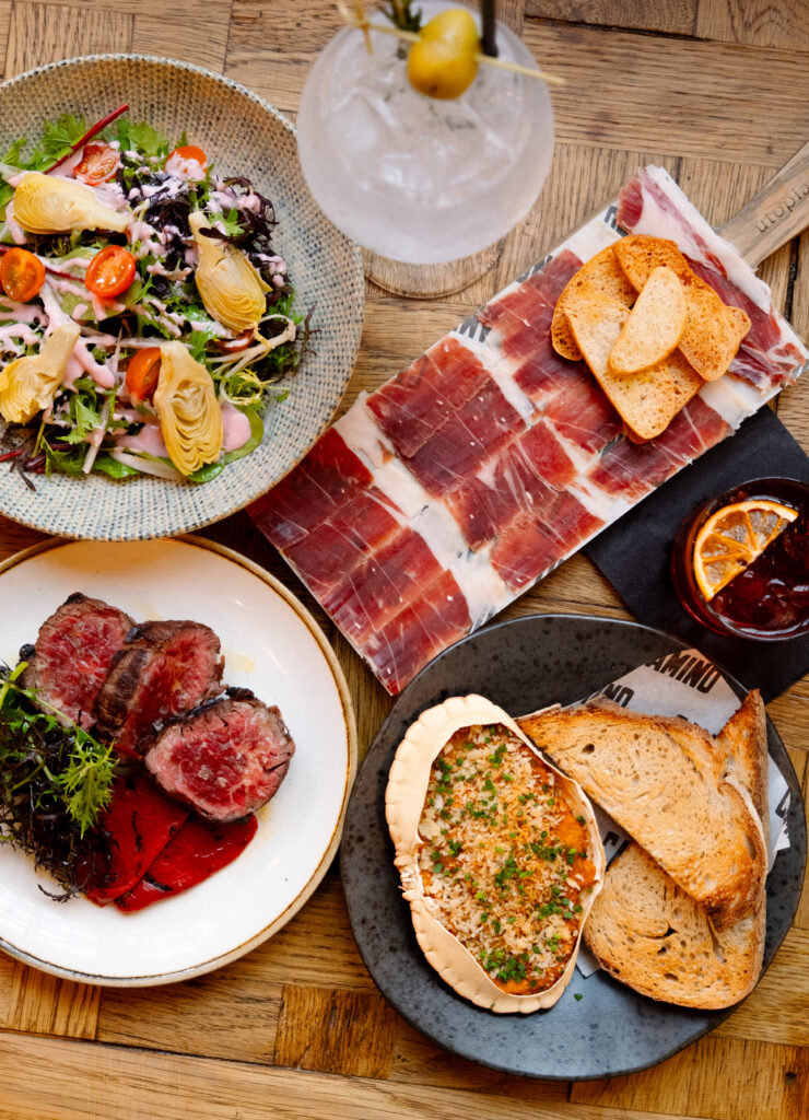 A spread of dishes including a salad with cherry tomatoes and artichokes, a plate of sliced cured meat with bread, sliced steak on a bed of greens, and a cheesy baked dish with toast. A cocktail with an olive is in the background.