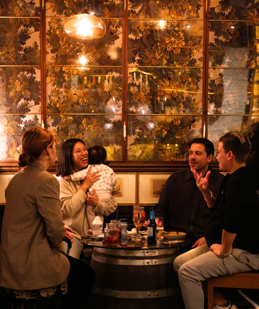 A group of five people, including one holding a baby, sit around a table in a cozy setting. They appear to be conversing and enjoying drinks. The wall behind them has a nature-themed design and warm lighting.