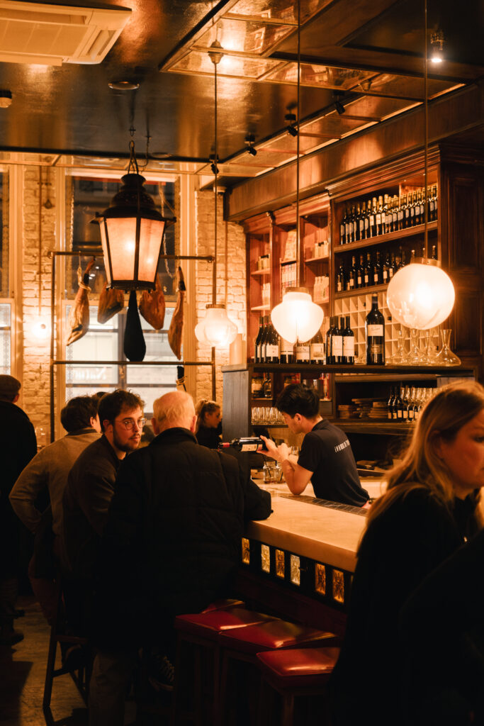 A warm-lit, cozy bar with wooden decor, hanging lamps, and shelves of wine bottles. Patrons sit at the counter, engaged in conversation, while a bartender serves drinks. The atmosphere is lively and intimate.