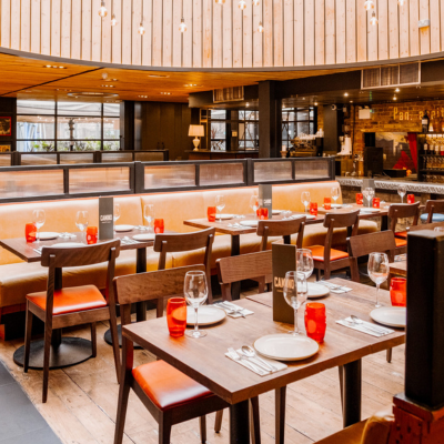 A restaurant interior with wooden tables and chairs featuring red cushions. Tables are set with white plates, cutlery, and red glasses. The warm lighting, wooden accents, and curved ceiling create a cozy ambiance. A bar area is visible in the background.