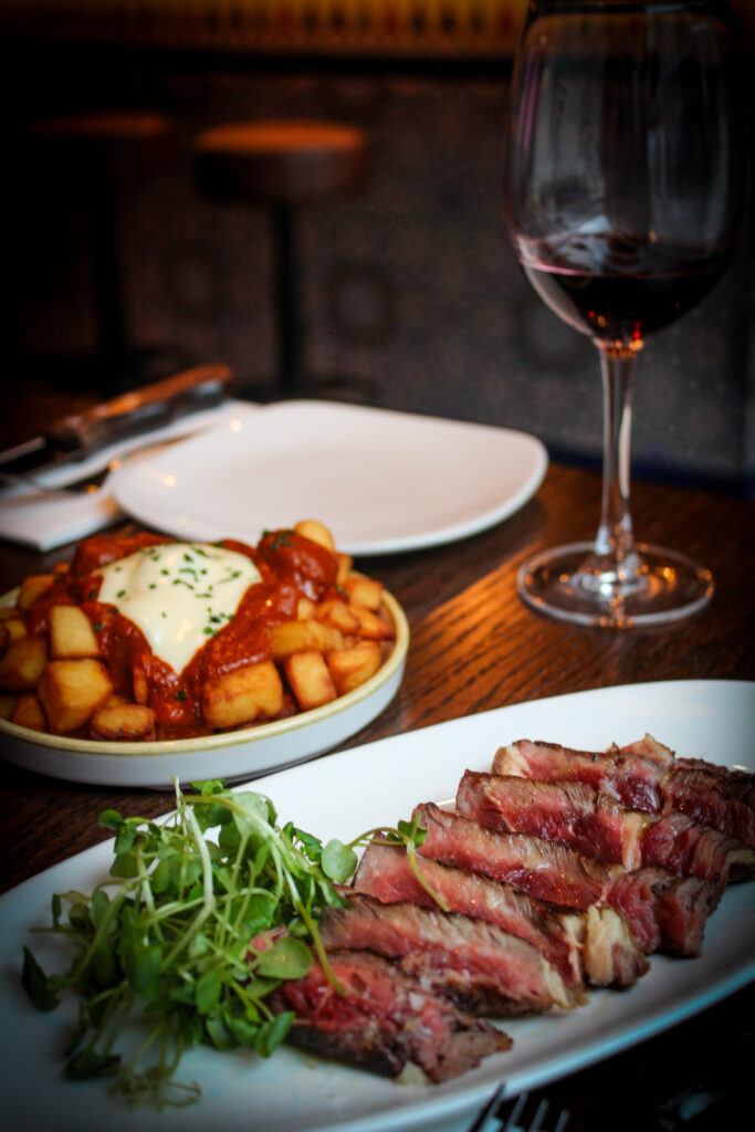 A plate of sliced rare steak with greens, a bowl of patatas bravas topped with aioli, and a glass of red wine on a wooden table in a restaurant setting.