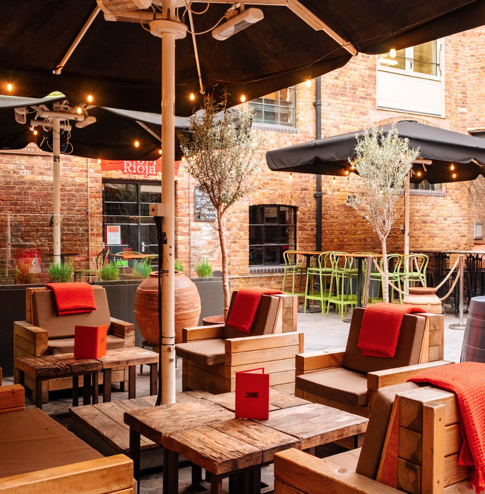 Outdoor patio with wooden tables and cushioned chairs, each with a red blanket and menu. String lights and large umbrellas provide shade. Green bar stools line a counter against a brick wall in the background.