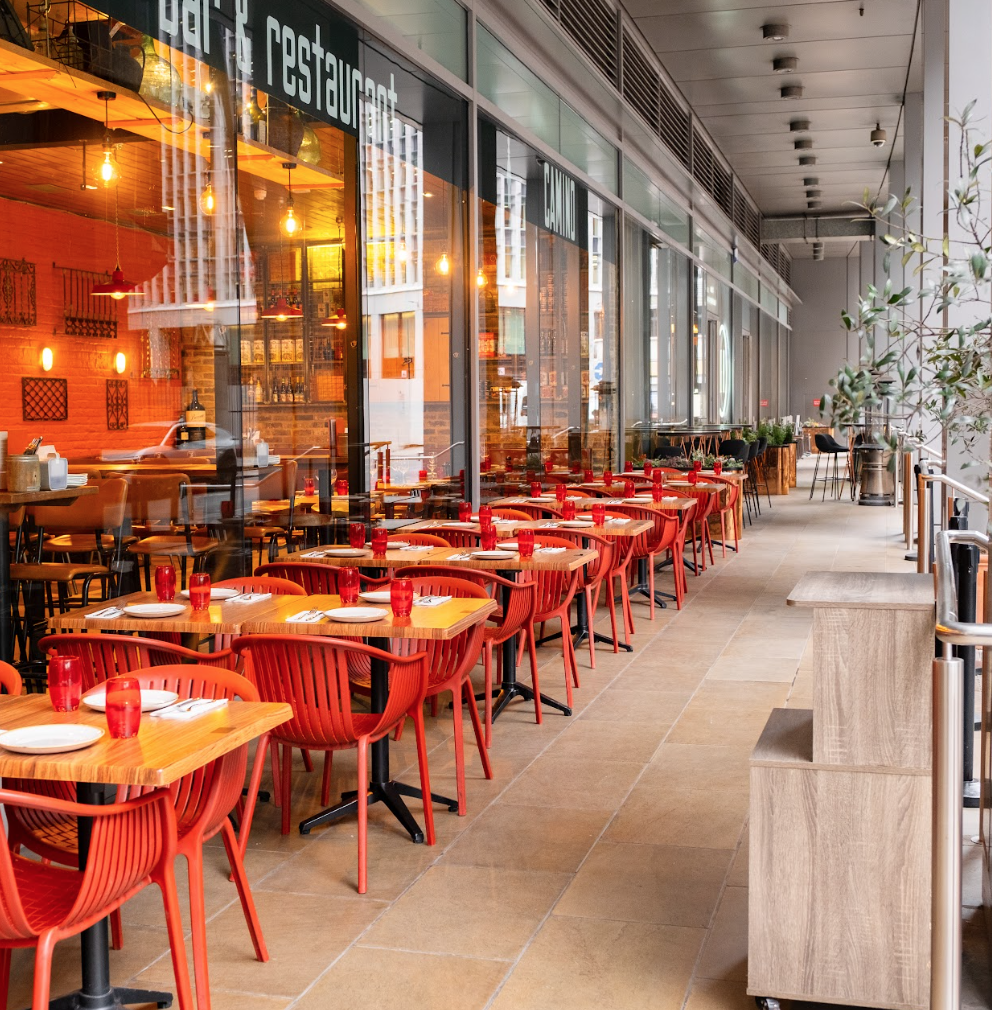 Outdoor restaurant seating area with red chairs and wooden tables set with plates and red glasses, next to large glass windows showing the indoor dining area with warm lighting.