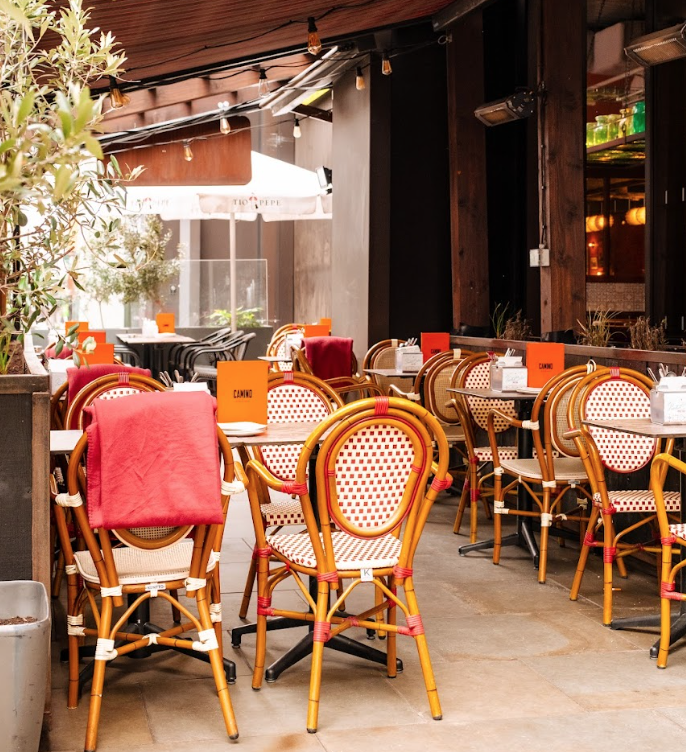 Outdoor café with round tables and rattan chairs, some draped with red blankets. Orange menu cards sit on each table. The area is shaded and cozy, with string lights hanging above.