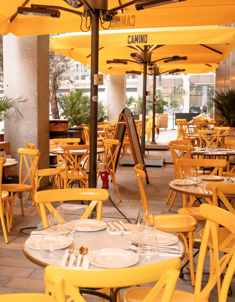 Outdoor cafe with yellow chairs and tables set for dining, beneath large yellow umbrellas. The scene is bright and inviting, with potted plants and a city setting in the background.