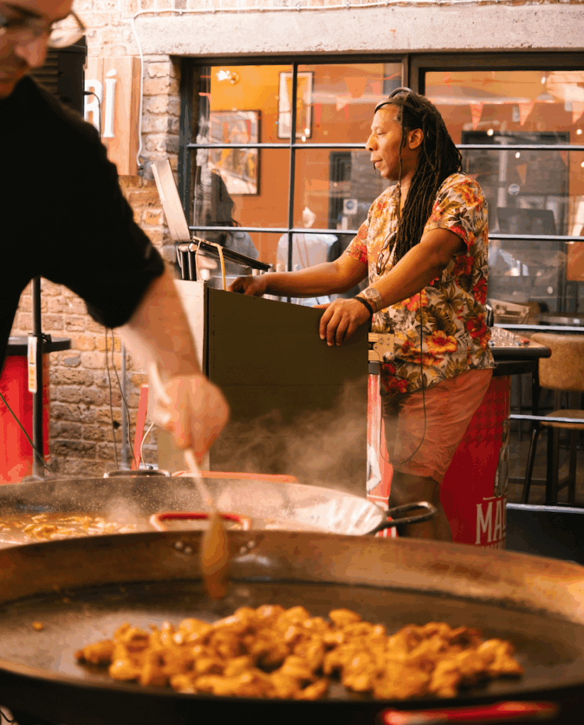 A man in a colorful floral shirt stands behind a DJ booth, while another person stirs food in a large pan in the foreground at an indoor event. Steam rises from the pan, and warm lighting fills the space.