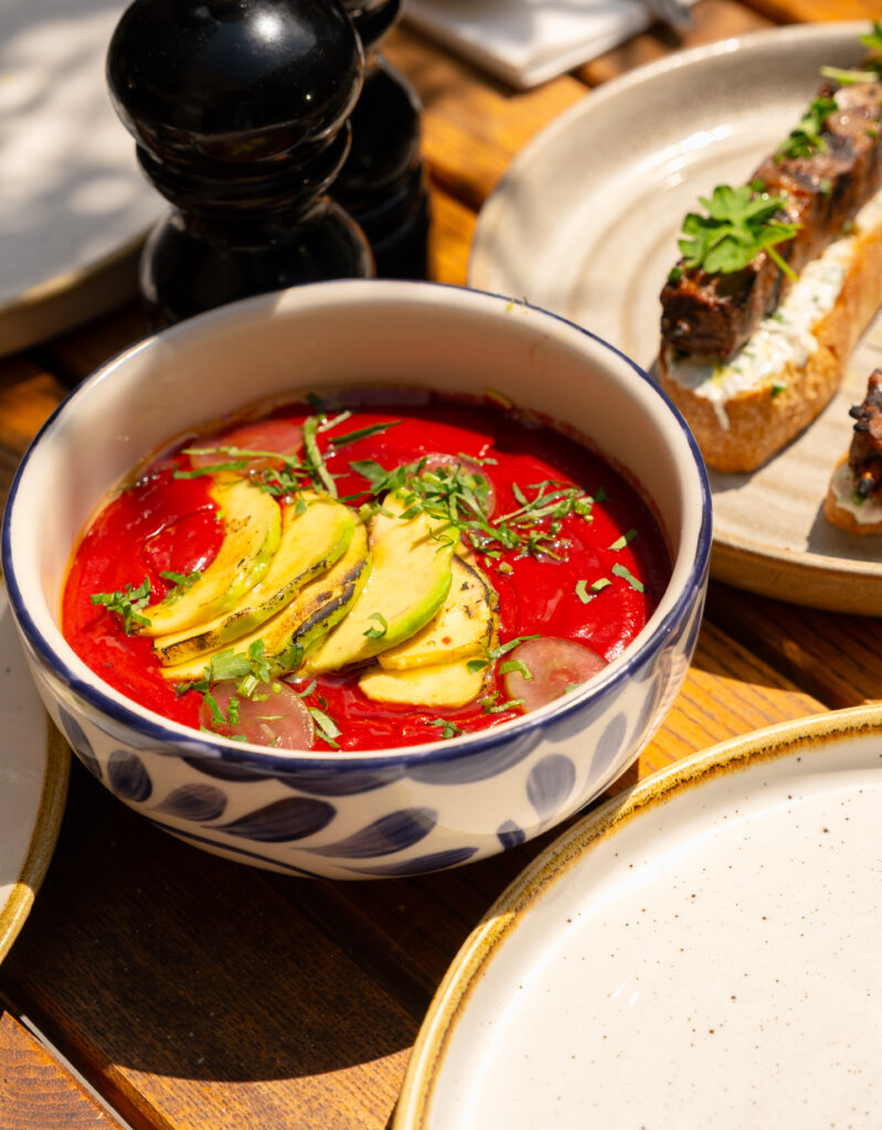 A bowl of vibrant red soup topped with sliced avocado, herbs, and small onions sits on a wooden table next to plates of food and black pepper shakers, bathed in warm sunlight.