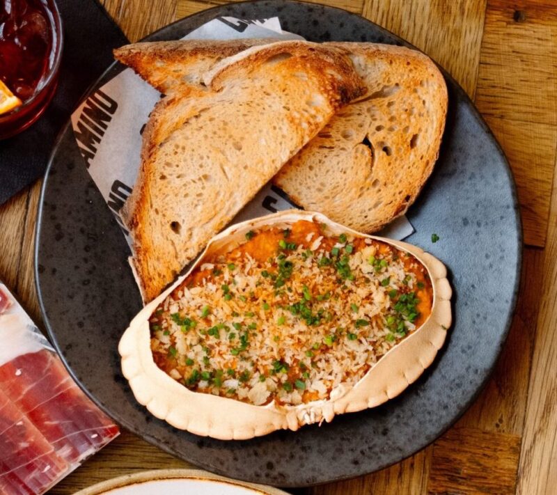 A plate with two slices of toasted bread and a crab shell filled with a baked seafood mixture, topped with breadcrumbs and chopped herbs, on a wooden table.