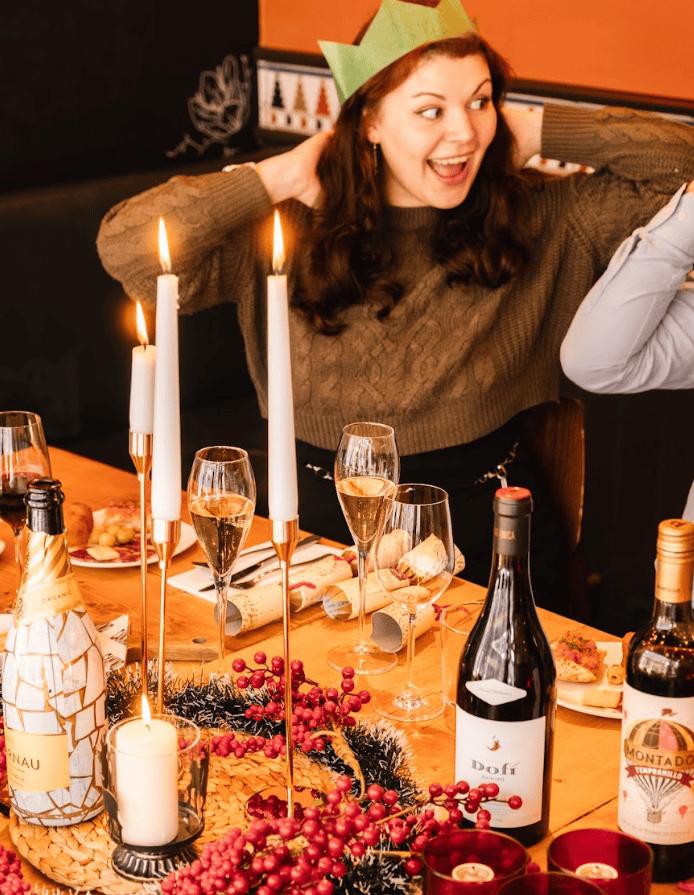 A smiling woman in a green paper crown sits at a festive table set with candles, wine, champagne, red berries, and food, enjoying a cheerful holiday celebration.