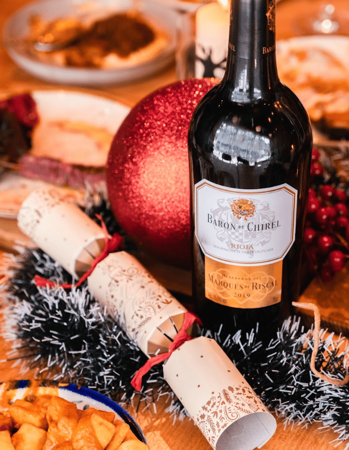 A bottle of Baron de Chirel red wine sits on a festive table with a Christmas cracker, black and white garland, a large red ornament, and plates of food in the background.