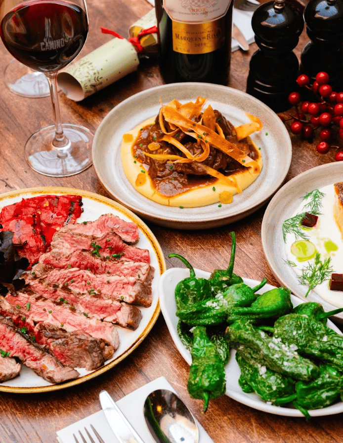 A table set with plates of grilled steak, roasted red peppers, sautéed green peppers, pasta with beef ragout, a glass and bottle of red wine, salt and pepper shakers, and festive holiday decorations.