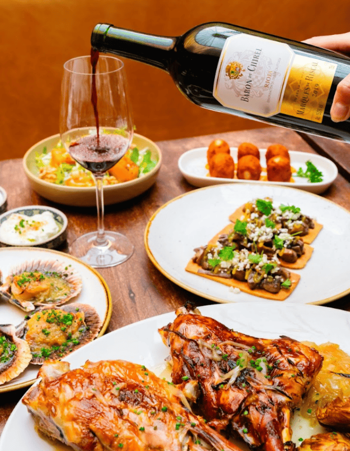 A hand pours red wine into a glass next to plates of roasted meat, scallops, flatbread with toppings, croquettes, and side dishes on a wooden table.