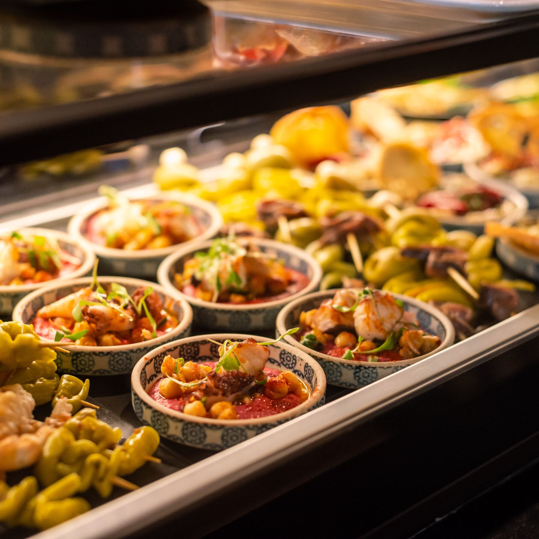 Small colorful bowls filled with various tapas, including shrimp, vegetables, and herbs, displayed in a glass case at a restaurant, with skewered appetizers in the background.