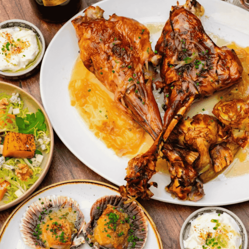 A plate with two roasted lamb shanks on a bed of sauce, surrounded by roasted mushrooms. Nearby are dishes with salad, baked scallops, and bowls of cream topped with herbs.
