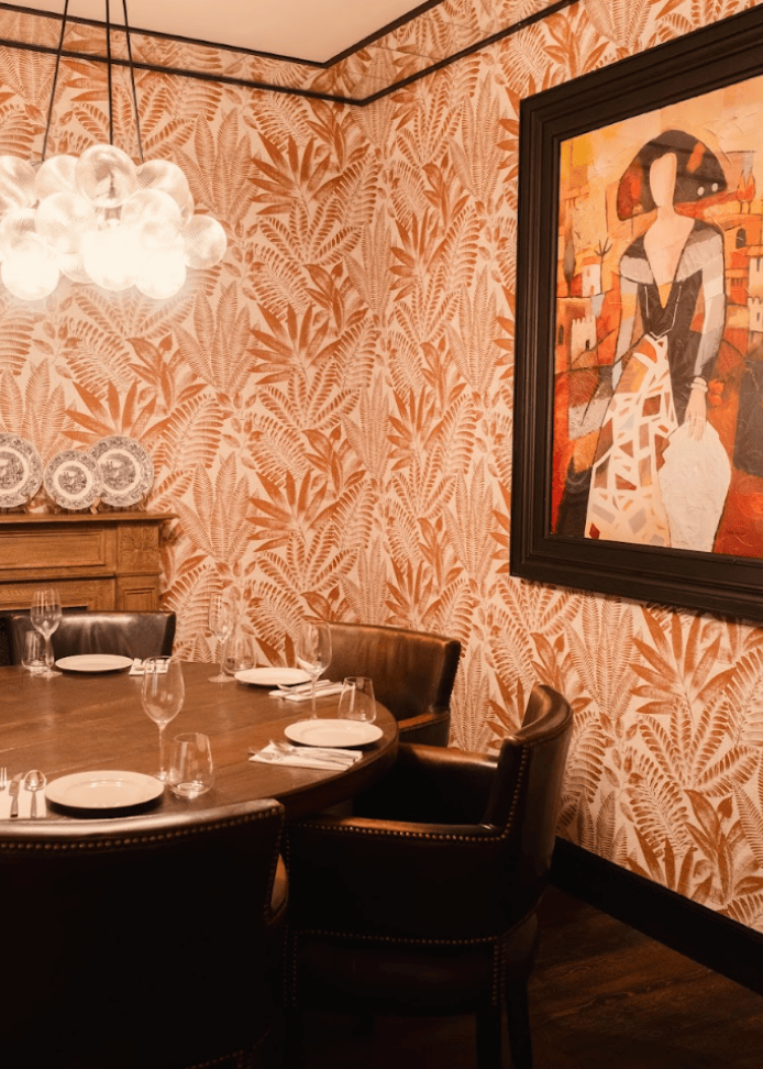 A dining room with a round table set for five, brown leather chairs, patterned wallpaper with leaves, a modern chandelier, decorative plates on a sideboard, and a large framed painting of a woman on the wall.