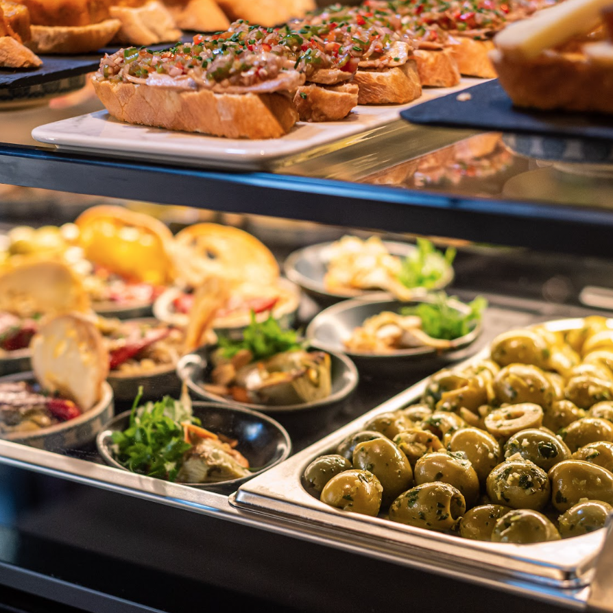 A display case filled with assorted tapas, including green olives and small plates with appetizers, and slices of bread with toppings arranged on upper shelves.