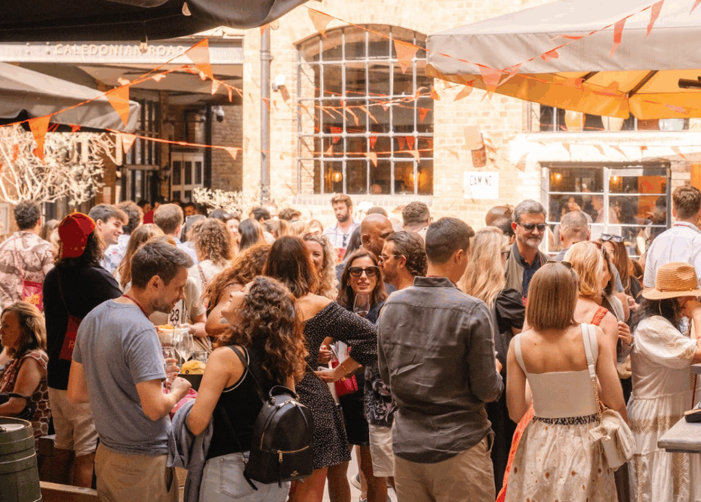 A lively crowd of people gathers outdoors under large umbrellas, socializing at a daytime event with orange bunting decorations and a brick building in the background.
