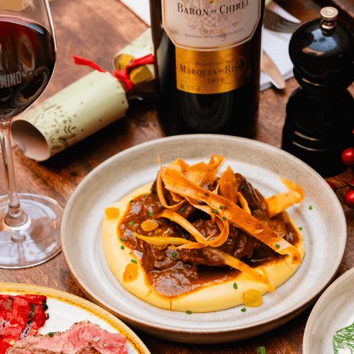 A plate of braised meat in rich sauce served over creamy mashed potatoes, garnished with fried parsnip strips. A bottle of red wine, a wine glass, and a pepper grinder are on the wooden table nearby.