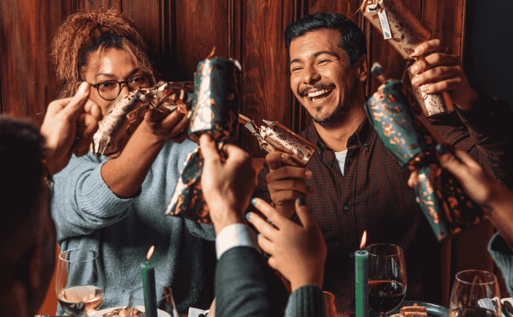 A group of people gathered around a table, smiling and pulling festive Christmas crackers together, celebrating joyfully with lit candles and wine glasses in the foreground.