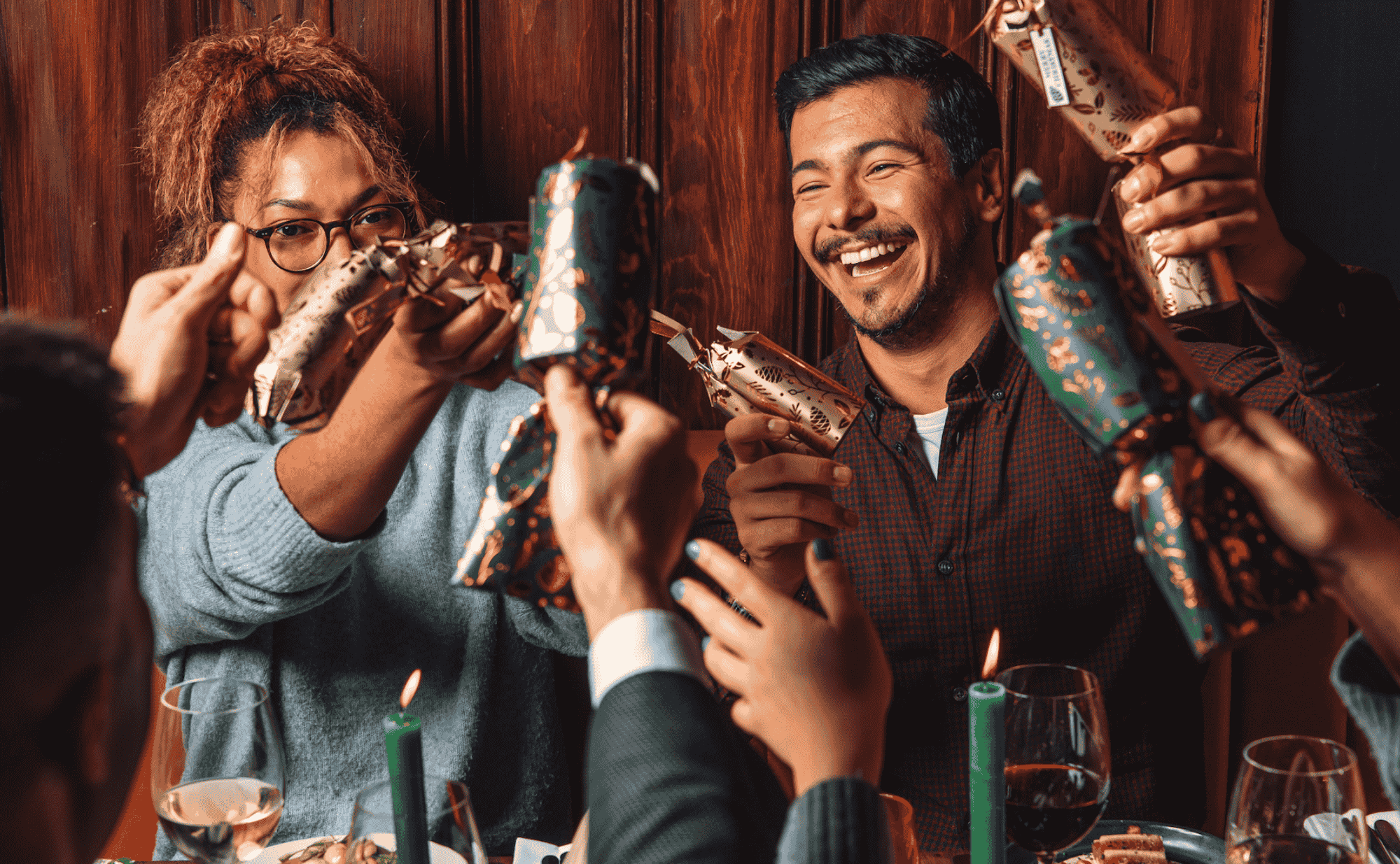 A group of people gathered around a table, smiling and pulling festive Christmas crackers together, celebrating joyfully with lit candles and wine glasses in the foreground.