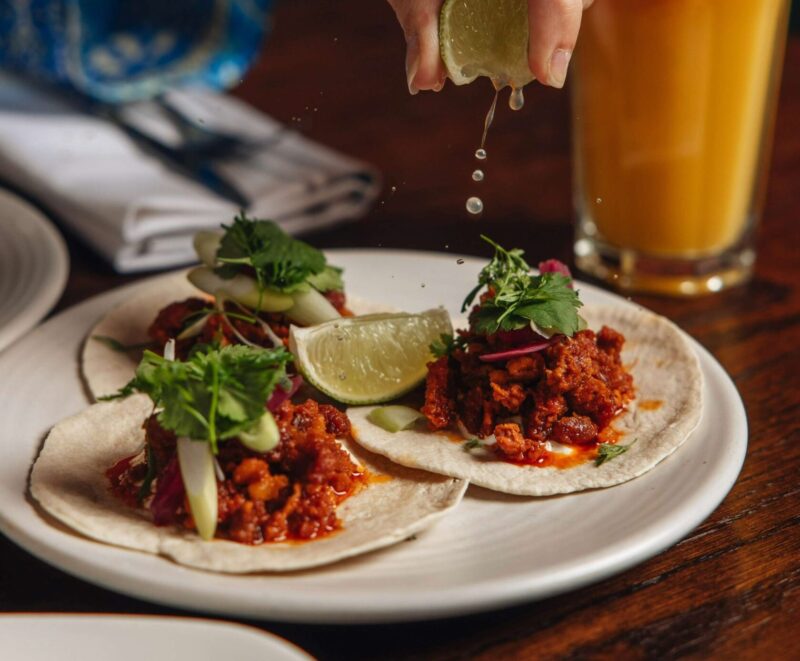 A hand squeezes a lime wedge over three tacos filled with seasoned meat, onions, and cilantro on corn tortillas, served on a white plate with a glass of orange drink in the background.