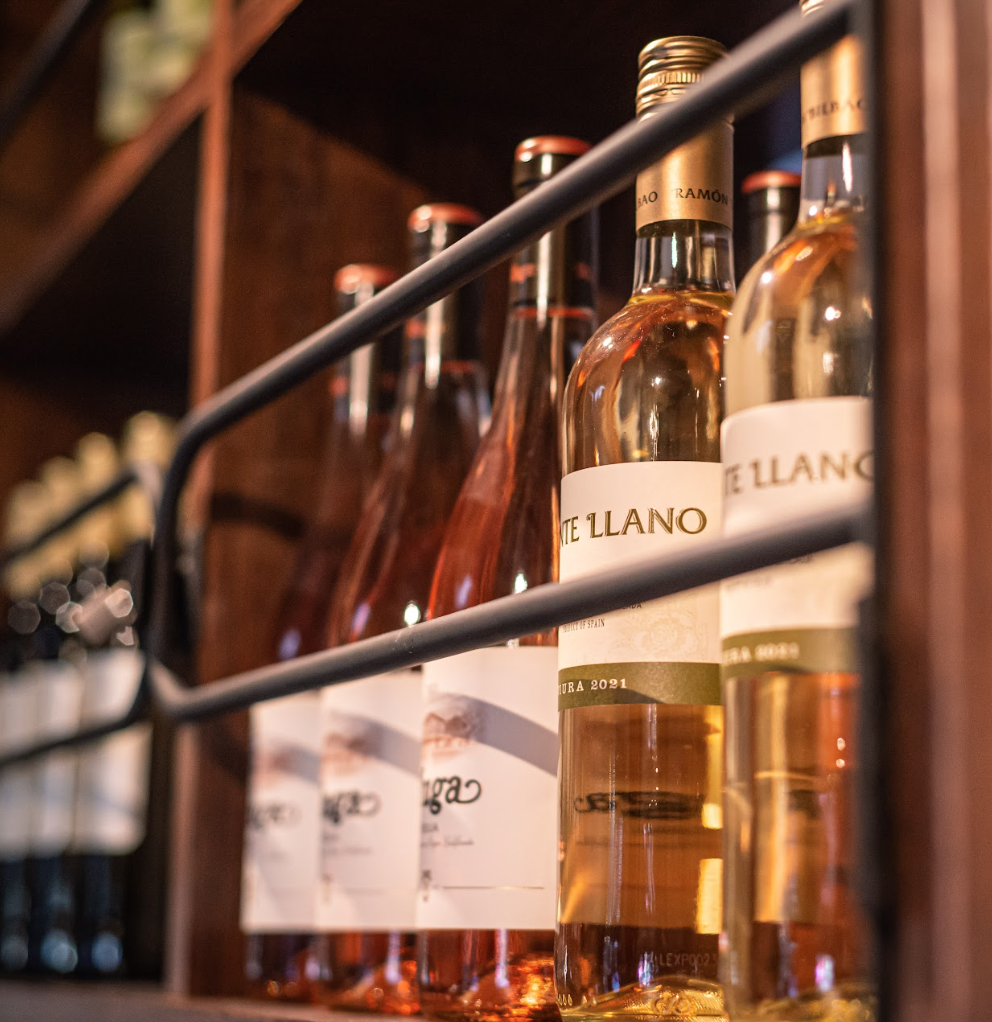 Bottles of wine, including rosé and white varieties, are neatly arranged on a wooden shelf behind a metal rail in a wine shop or cellar.