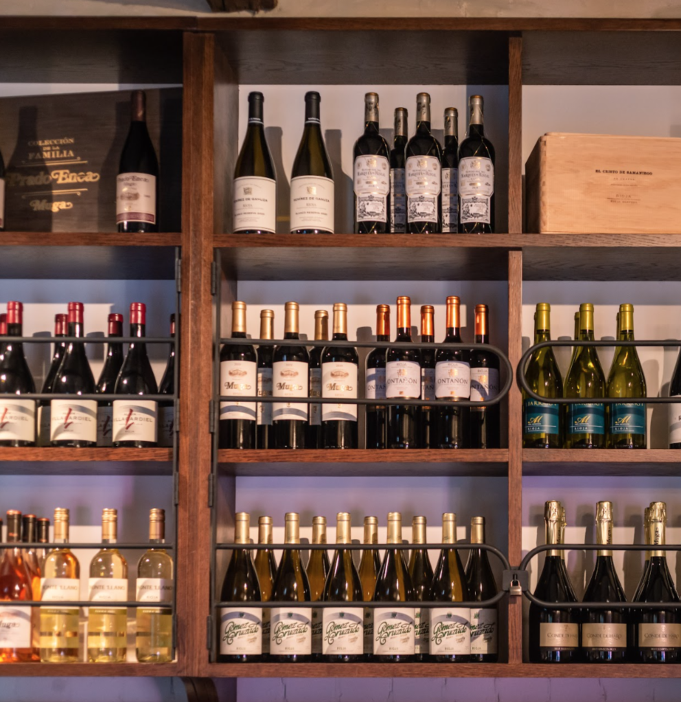 A wooden shelf displays various bottles of wine, arranged by type and label, with some bottles stacked upright and others tilted horizontally. A wooden box and wine accessories are also visible on the shelves.