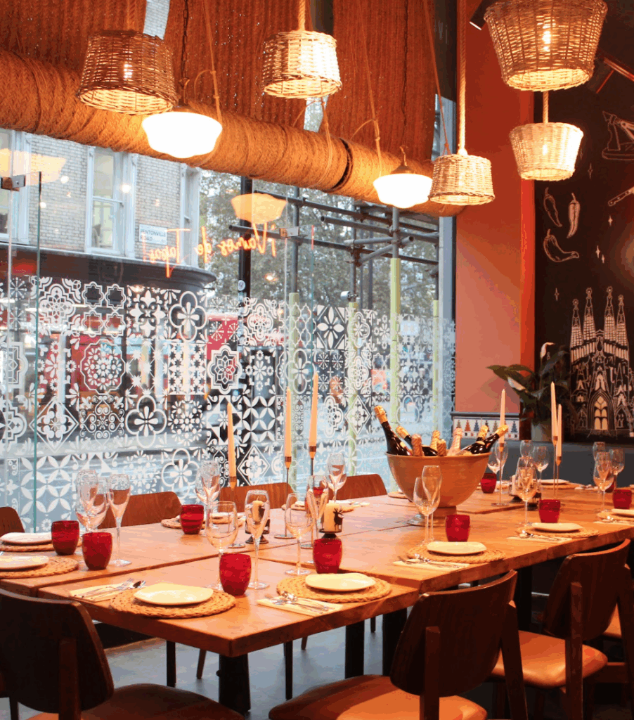 A long wooden dining table set for a meal with red glass cups, wine glasses, and candles, surrounded by modern wooden chairs under hanging woven lamps in a stylish restaurant with large windows and patterned glass.