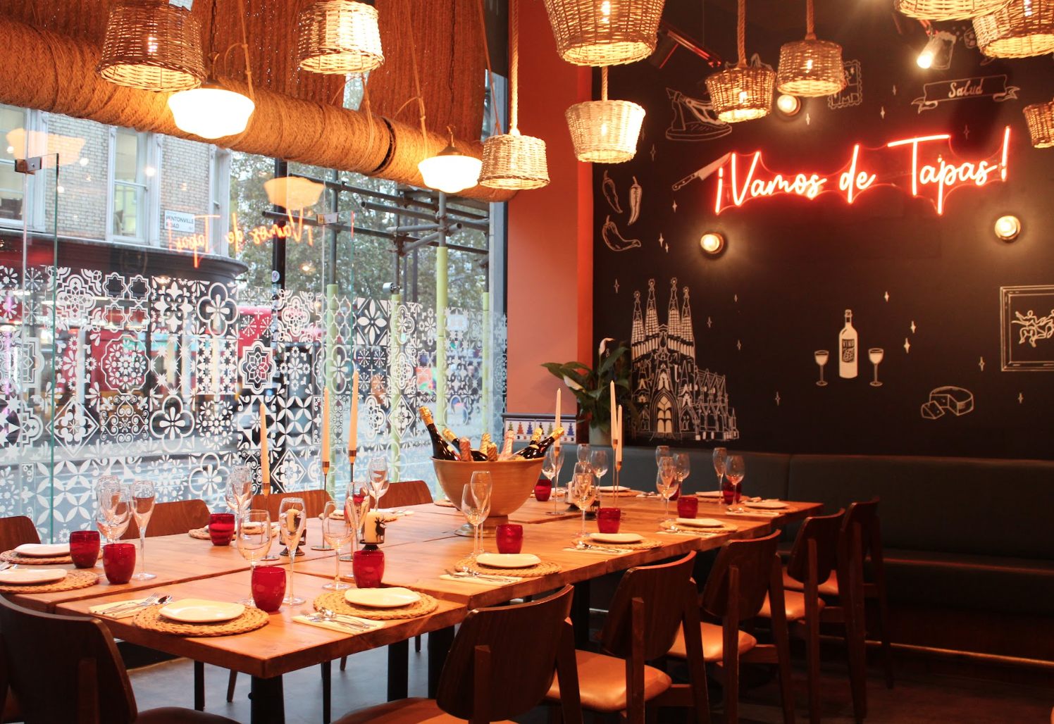 A cozy restaurant with a long wooden table set for dining, wicker pendant lights, decorative wall art, and a neon sign reading 