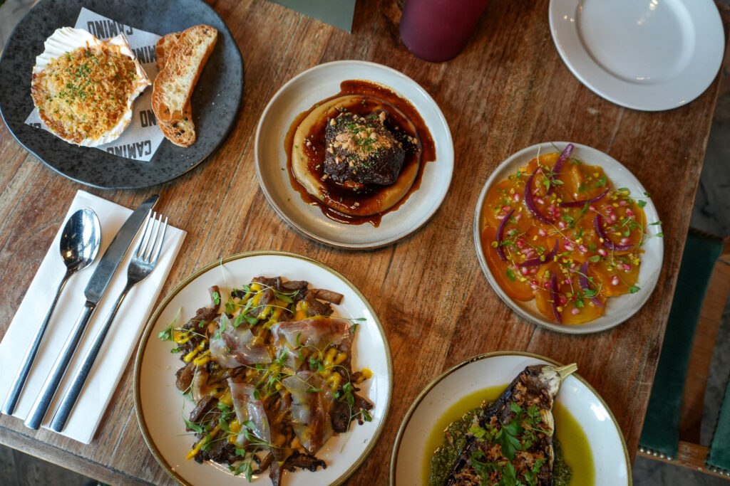 Five plates of assorted gourmet dishes, including bread with spread, grilled meat, marinated vegetables, and a colorful salad, are arranged on a rustic wooden table with cutlery and a napkin beside them.
