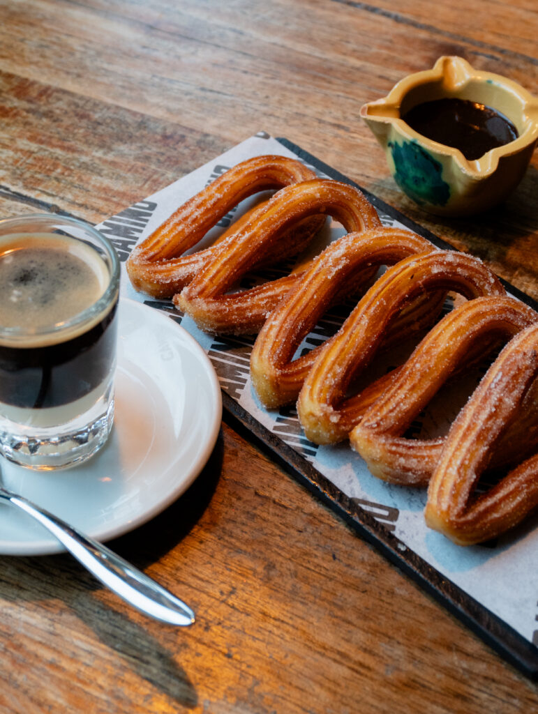 A plate of four churros dusted with sugar, served with a small cup of chocolate sauce and a glass of espresso on a wooden table.