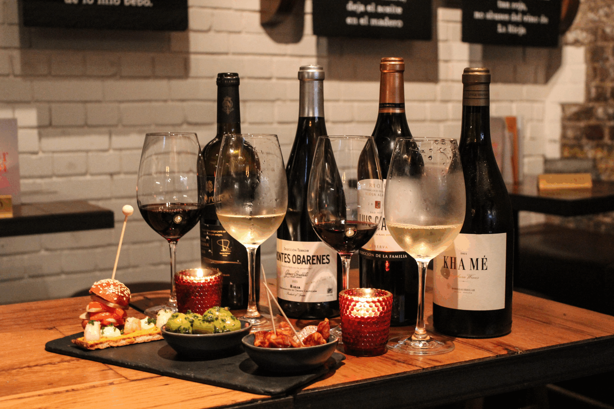 A wooden table with four bottles of wine, three filled wine glasses, and small plates of appetizers, including skewers, bruschetta, and olives, with red votive candles for ambiance.