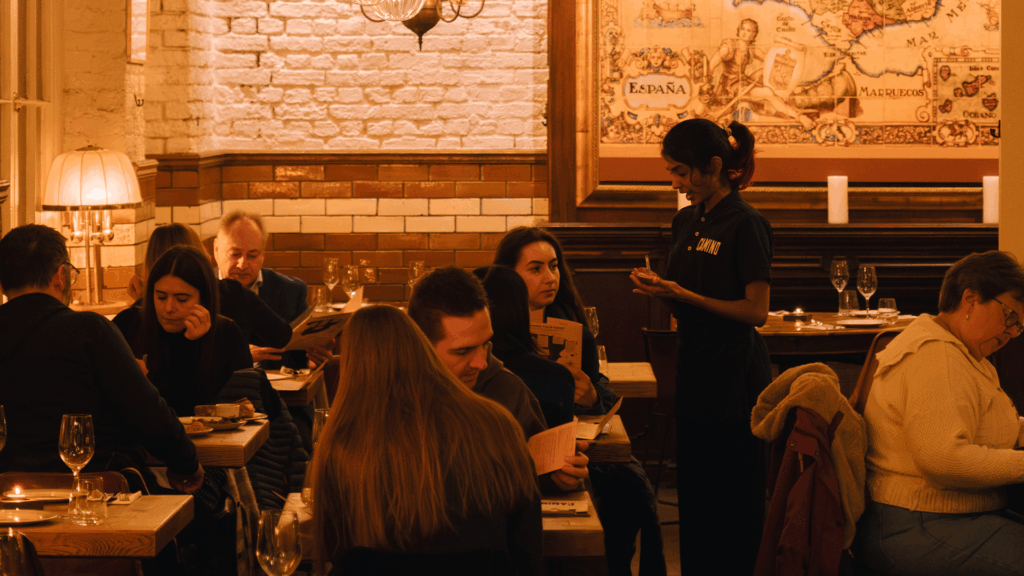 A warmly lit restaurant with diners sitting at tables, some reading menus and others conversing. A server stands taking an order. The walls are decorated with a large vintage map and classic lighting.