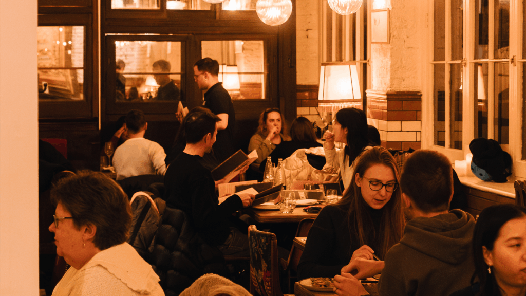 A cozy restaurant scene with several people sitting at tables, eating, drinking, and talking. Warm lighting creates a relaxed atmosphere, and some people are looking at menus while others are engaged in conversation.