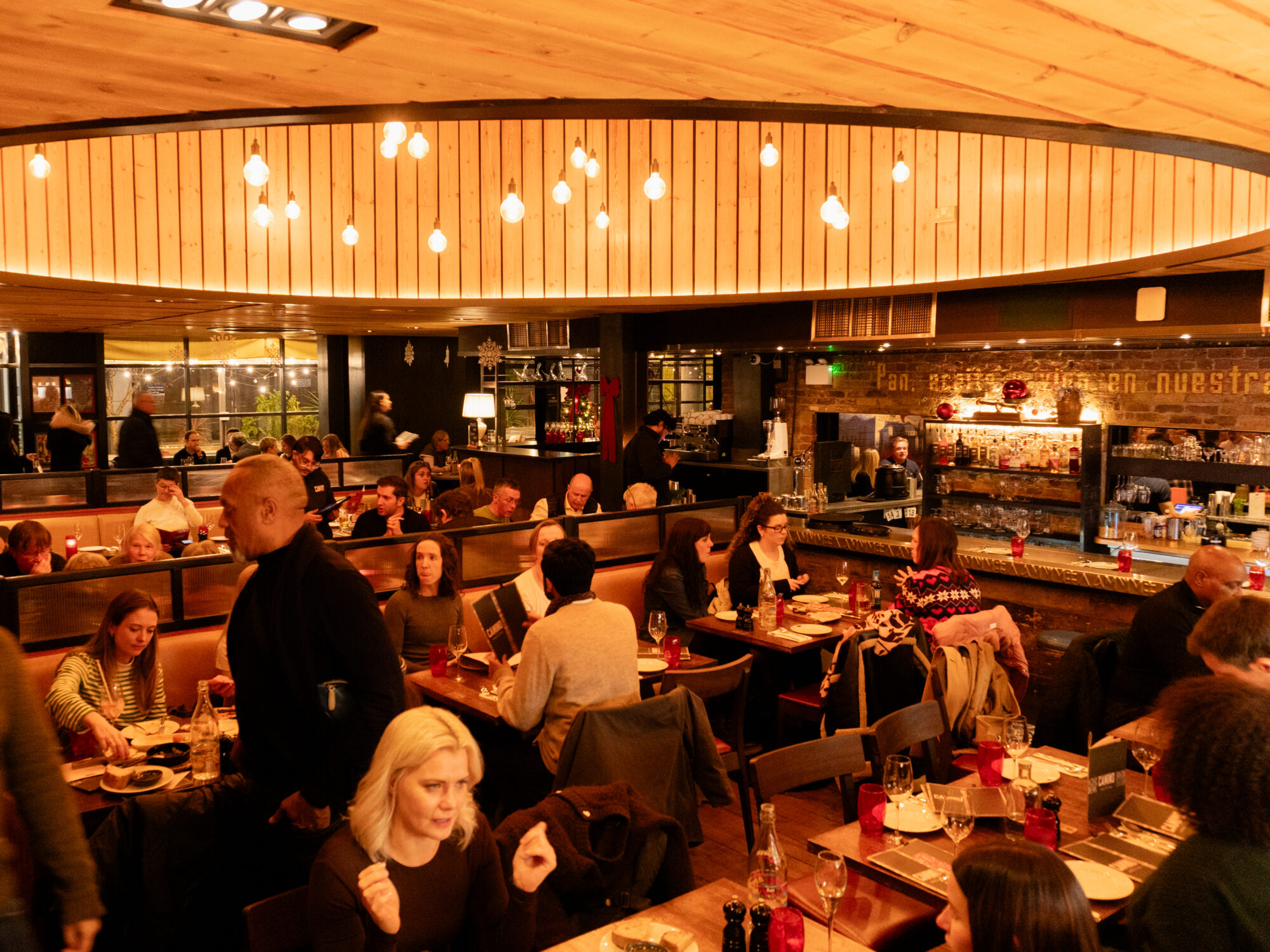 A busy, warmly lit restaurant filled with people dining and talking at wooden tables, with a central bar area and hanging lights creating a cozy atmosphere.