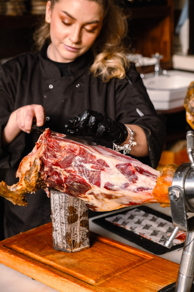 A chef wearing a black uniform and gloves slices a large cured ham leg mounted on a wooden stand in a kitchen setting.