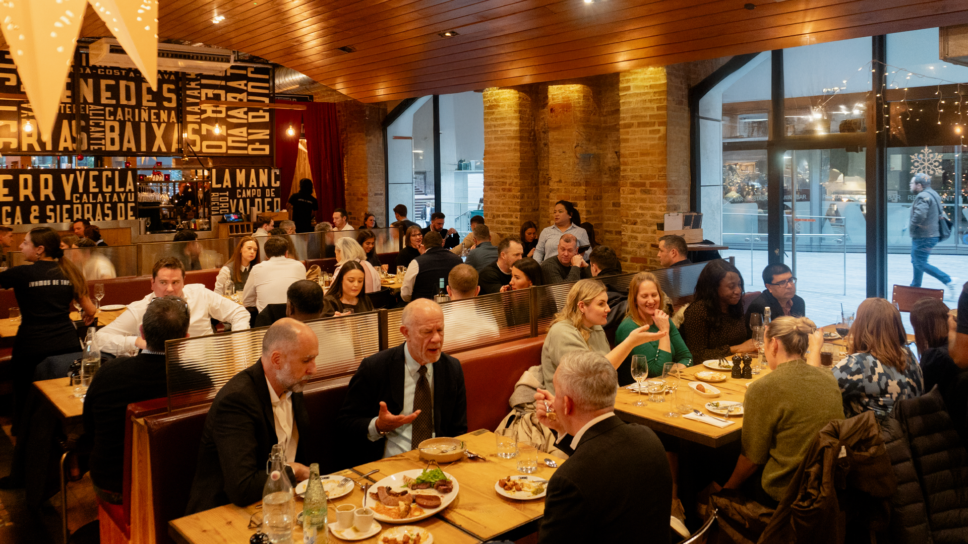 A busy restaurant with people sitting at wooden tables, eating, drinking, and talking. The atmosphere is lively and warm, with large windows and festive decorations visible outside.