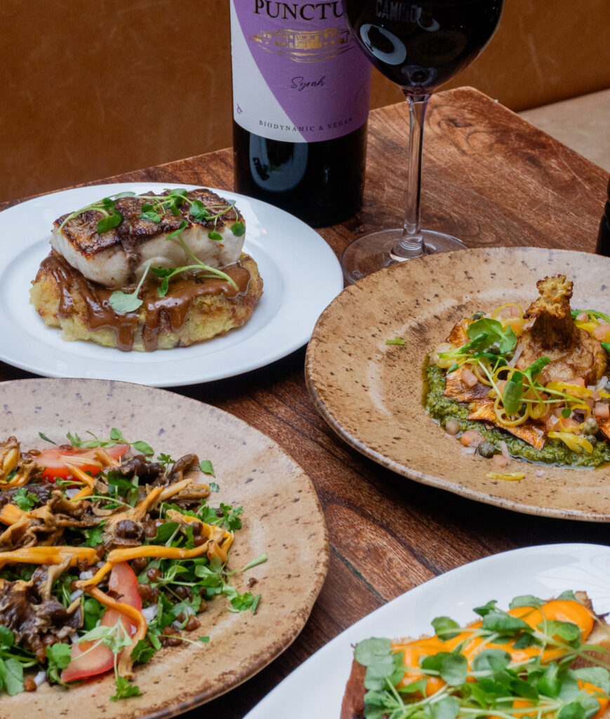 Four plates of gourmet dishes sit on a wooden table alongside a bottle of red wine and a glass. The dishes include fish on a fritter, salads with greens, sautéed mushrooms, and a colorful entrée with microgreens.
